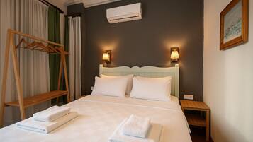A neatly made bed with two pillows and two folded towels, a wooden hanger, a wall-mounted air conditioner, and a bedside table with a lamp.