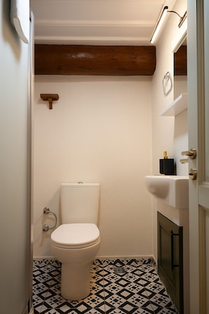 A small bathroom with a toilet, a wooden beam ceiling, a black and white patterned floor, and a white sink with a soap dispenser. - Defne Pansiyon Ayvalık (Ayvalık)
