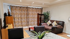 A living room with a sofa, a dining table with chairs, a potted plant, and a curtain with a leaf pattern. - Paramount Nest (Swindon)