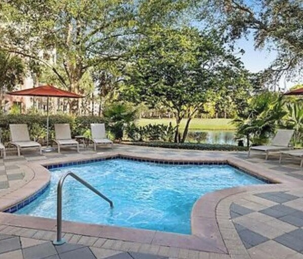 Una piscina con una escalera metálica, rodeada de sillas de descanso y una sombrilla roja.