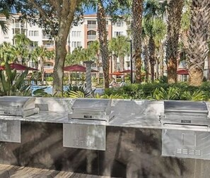 Zona junto a la piscina con una parrilla de metal y vista a un edificio de hotel al fondo.