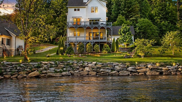 A multi-story house with a balcony, situated by a river, surrounded by trees and a stone wall. - Tower on the Tuck: Entire home on the Tuckaseegee River near WCU -Riverfront (Cullowhee)