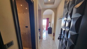 Un couloir avec un sol carrelé, une porte ornée d’un motif géométrique et une vue sur une pièce où l’on aperçoit une machine à laver et un rideau rouge.