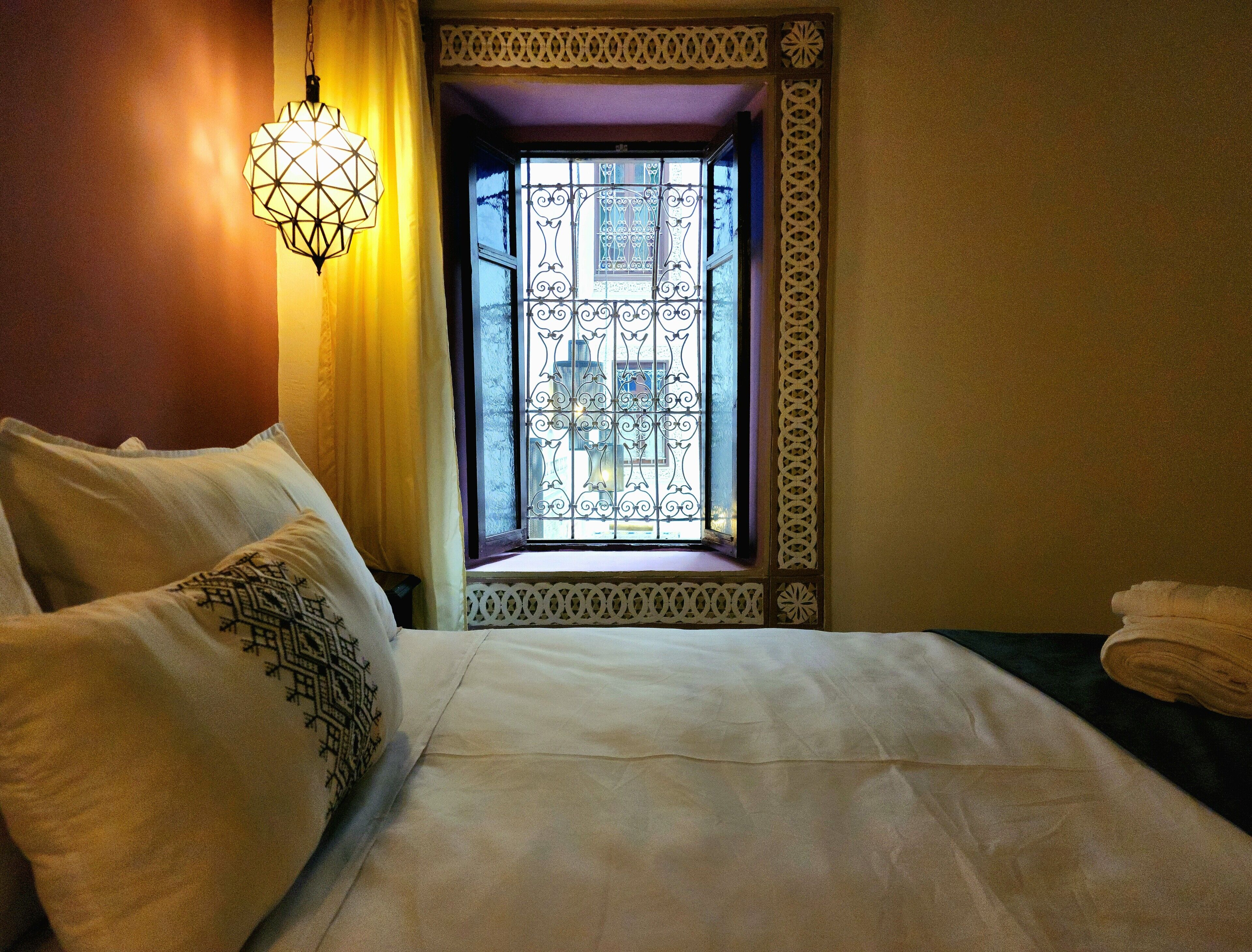 Una habitación de hotel con una cama, un ventanal decorativo con ornamentación metálica intrincada y una lámpara colgante.