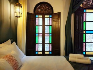 A hotel room with a bed, a nightstand, a wall-mounted lamp, and a window with stained glass. - Riad Dar Mazaj Fes (Fes)