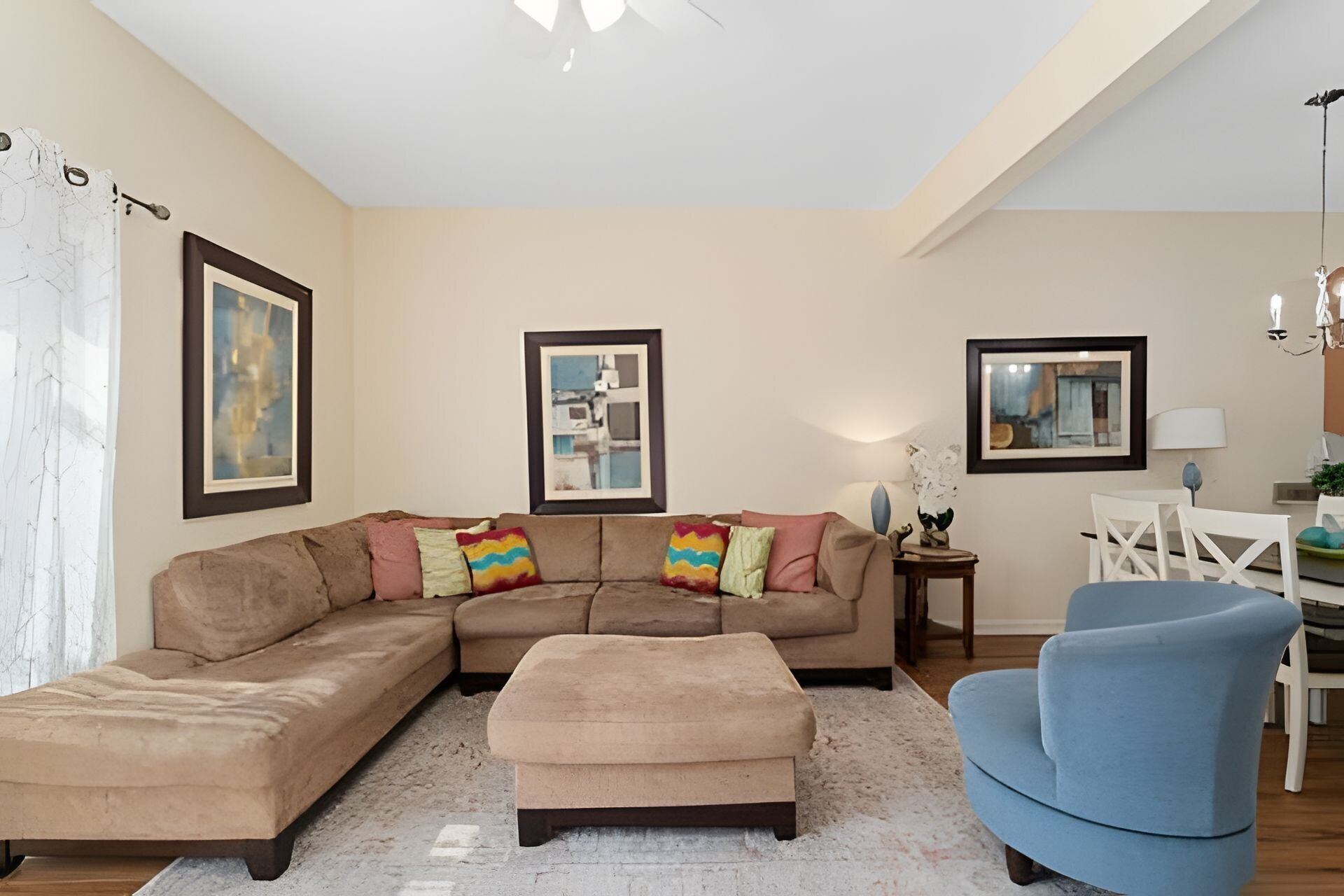 A living room with a sectional sofa, a blue armchair, and a small table with a lamp. There are two framed pictures on the walls.