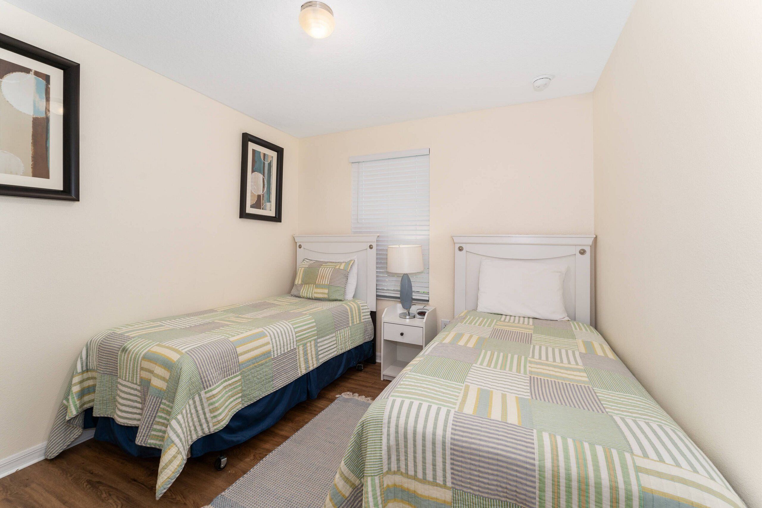 A bedroom with two single beds, a nightstand with a lamp, a framed picture on the wall, and a window with blinds.