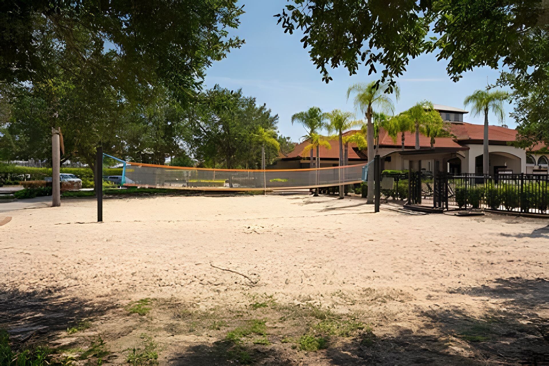 A sandy volleyball court with a net, surrounded by trees and a building with a red roof in the background.