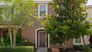 A two-story house with a brown facade, a front door, and a small garden with various plants and a tree. - Bella Vida Cozy 3bd/3ba King Bed Private Pool (Kissimmee)