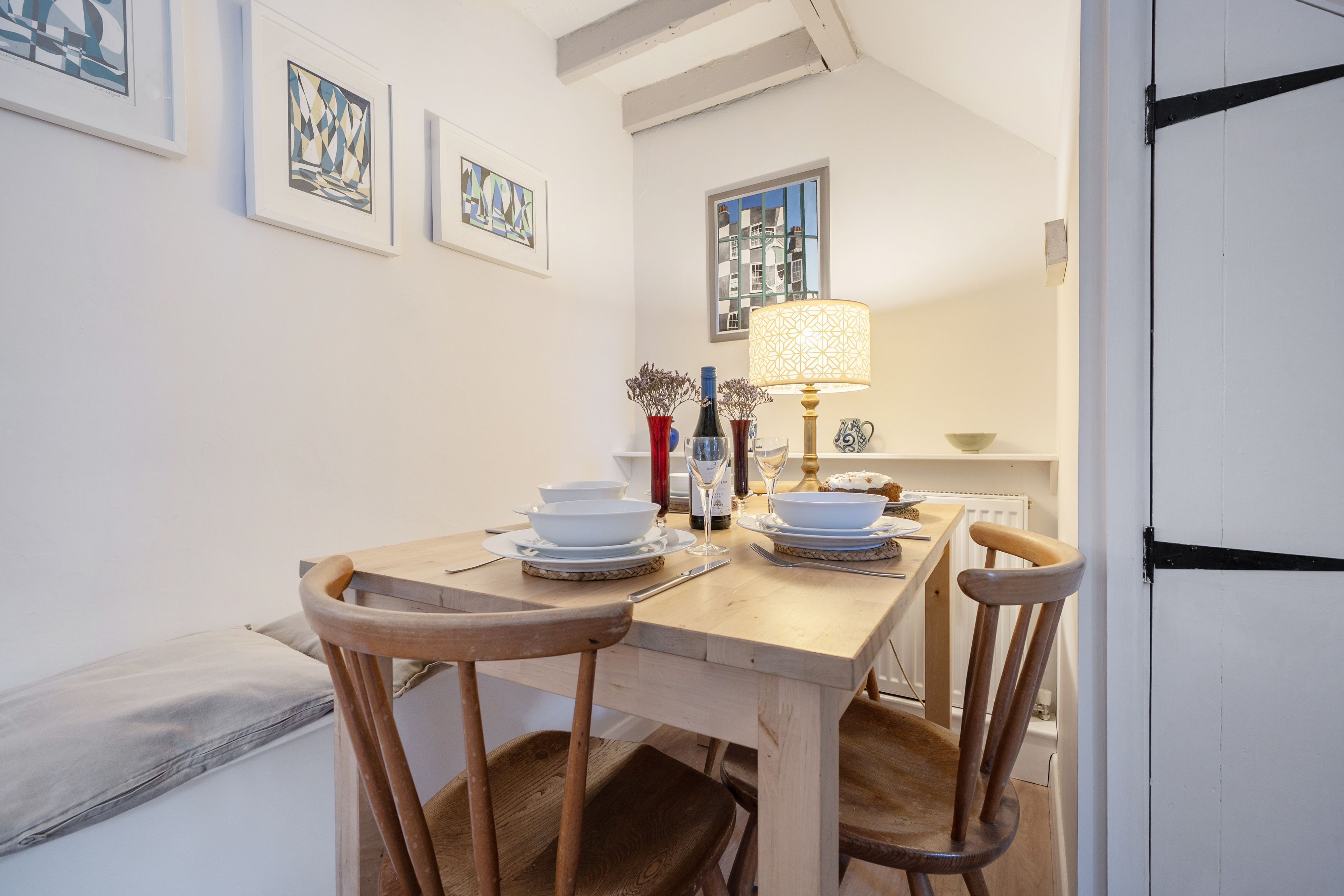 A dining table set with plates, bowls, and a lamp, surrounded by wooden chairs in a room with white walls and framed artwork.