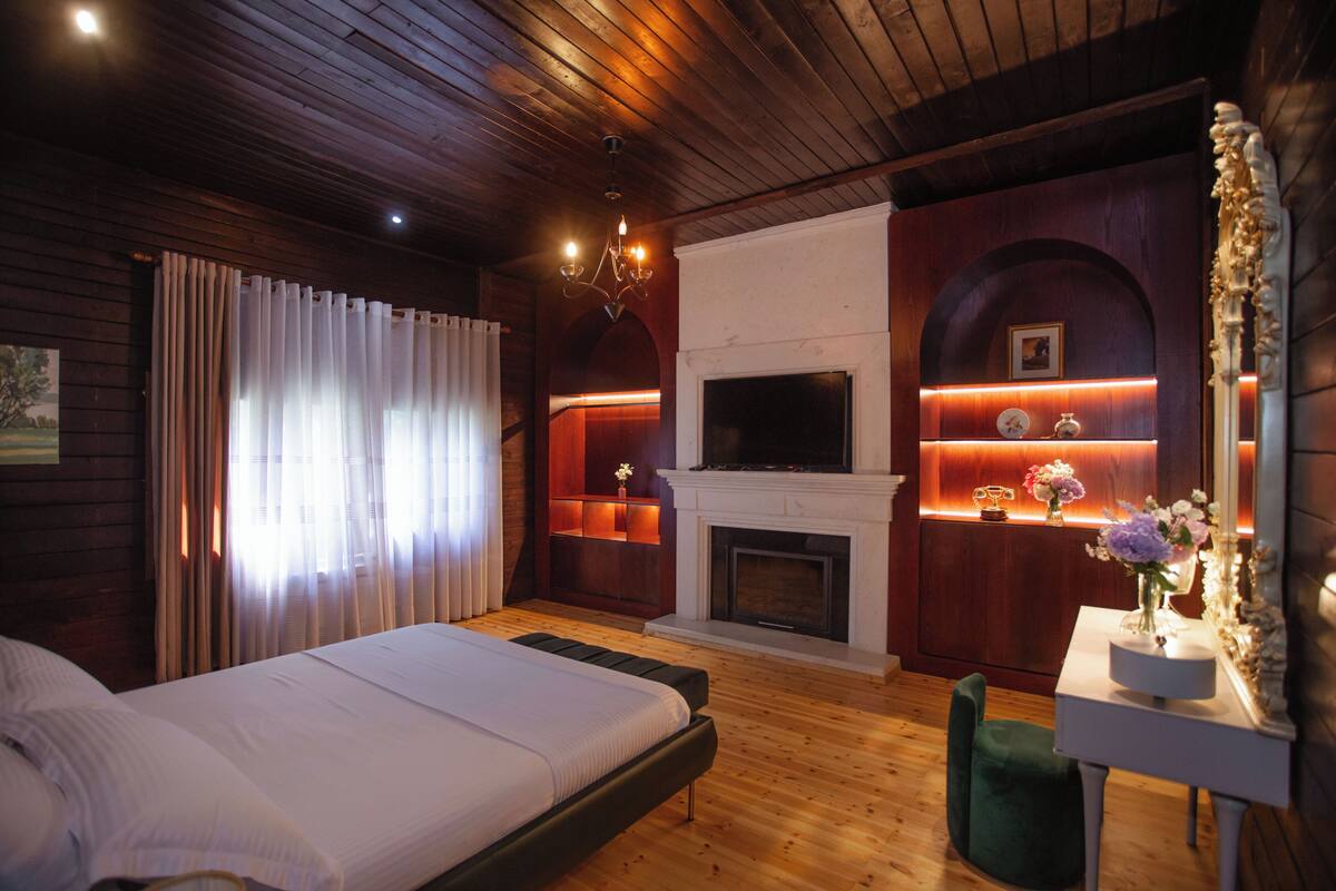 A bedroom with a bed, a fireplace, a TV, and a desk with a vase of flowers.
