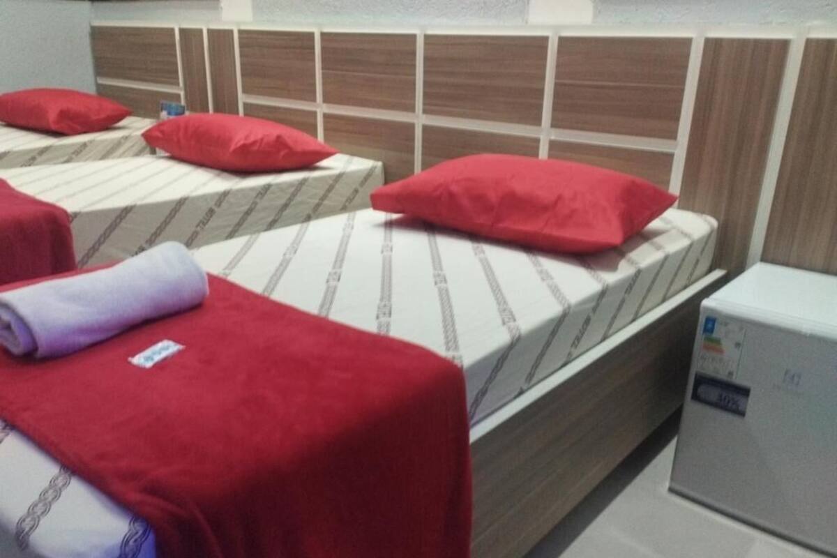 Habitación de hotel con dos camas, cada una con una almohada roja y un cubrecolchón a rayas blancas y grises. Se observa un electrodoméstico blanco pequeño a la derecha de la habitación.