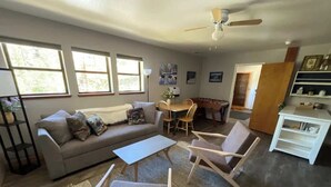 Ein Wohnzimmer mit einer Couch, zwei Sesseln, einem Couchtisch und einem Esstisch mit Stühlen. Es gibt Fenster, einen Deckenventilator und ein Bücherregal.