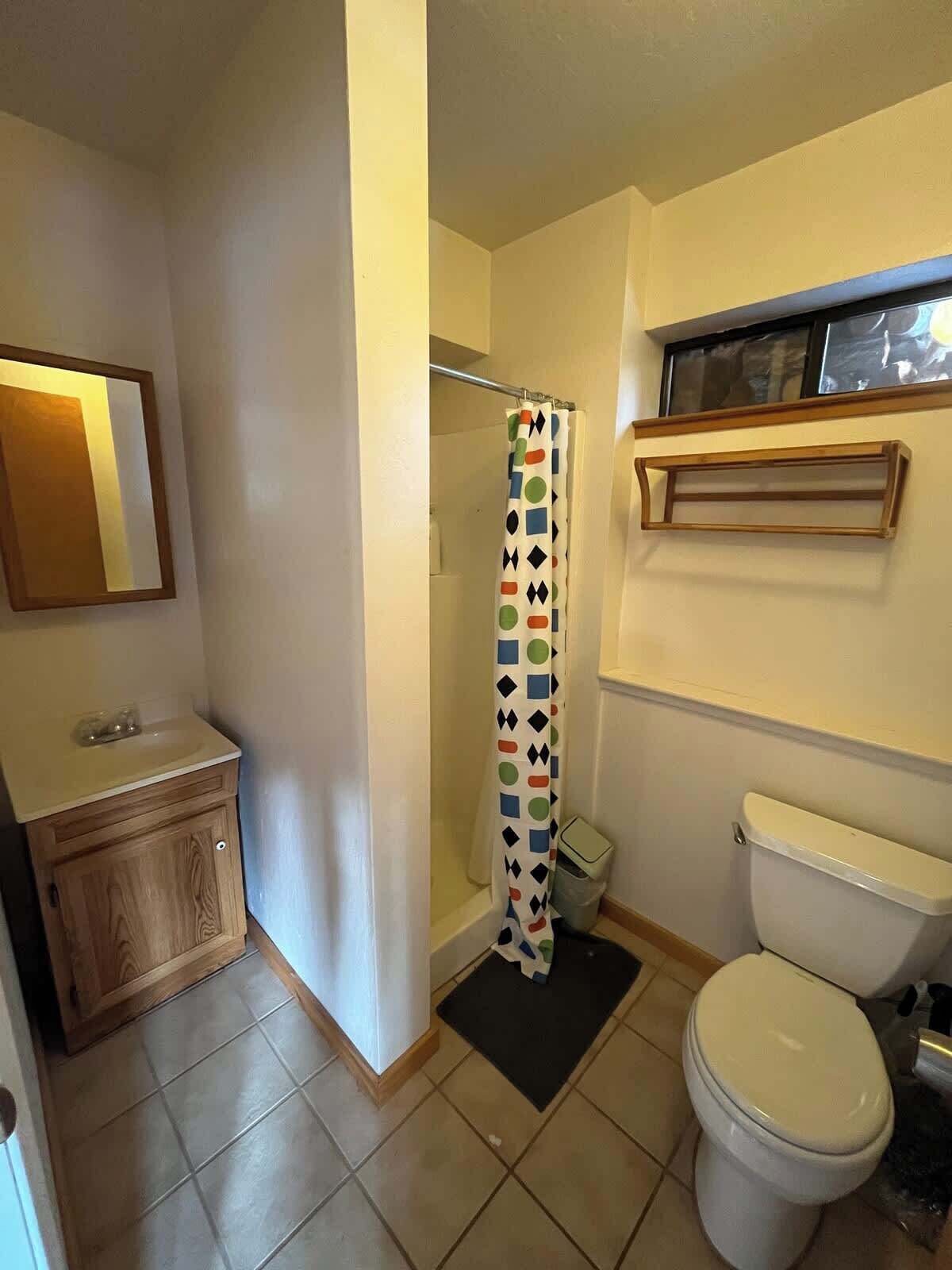 Une salle de bain comprenant des toilettes, un lavabo, un meuble en bois, un rideau de douche et une petite fenêtre située au-dessus des toilettes.