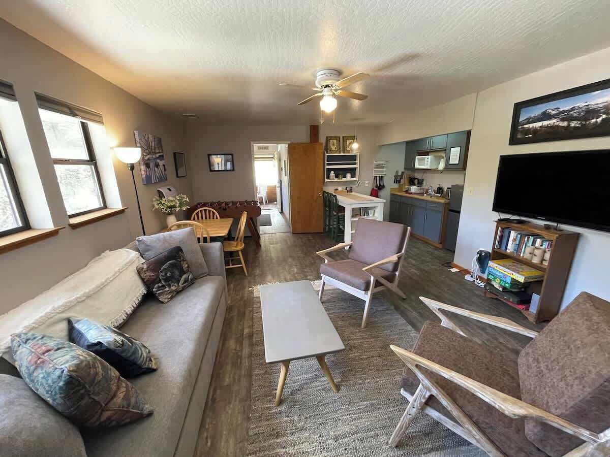 A living room with a sofa, armchairs, a coffee table, and a TV. There is a kitchen area in the background.