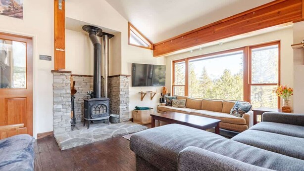A cozy living room with a stone fireplace, wooden beams, a sofa, and a large window offering a view of trees. - West Village-Large Lovely Private Home in Kirkwood (Kirkwood)