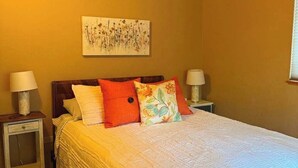 Een slaapkamer met een houten plafond, een bed met wit beddengoed en kleurrijke kussens, twee nachtkastjes met lampen en een bloemig kunstwerk aan de muur.