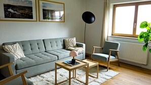 A living room with a grey sofa, a wooden coffee table, a blue armchair, and a floor lamp. There are two framed pictures on the wall and a window with curtains.