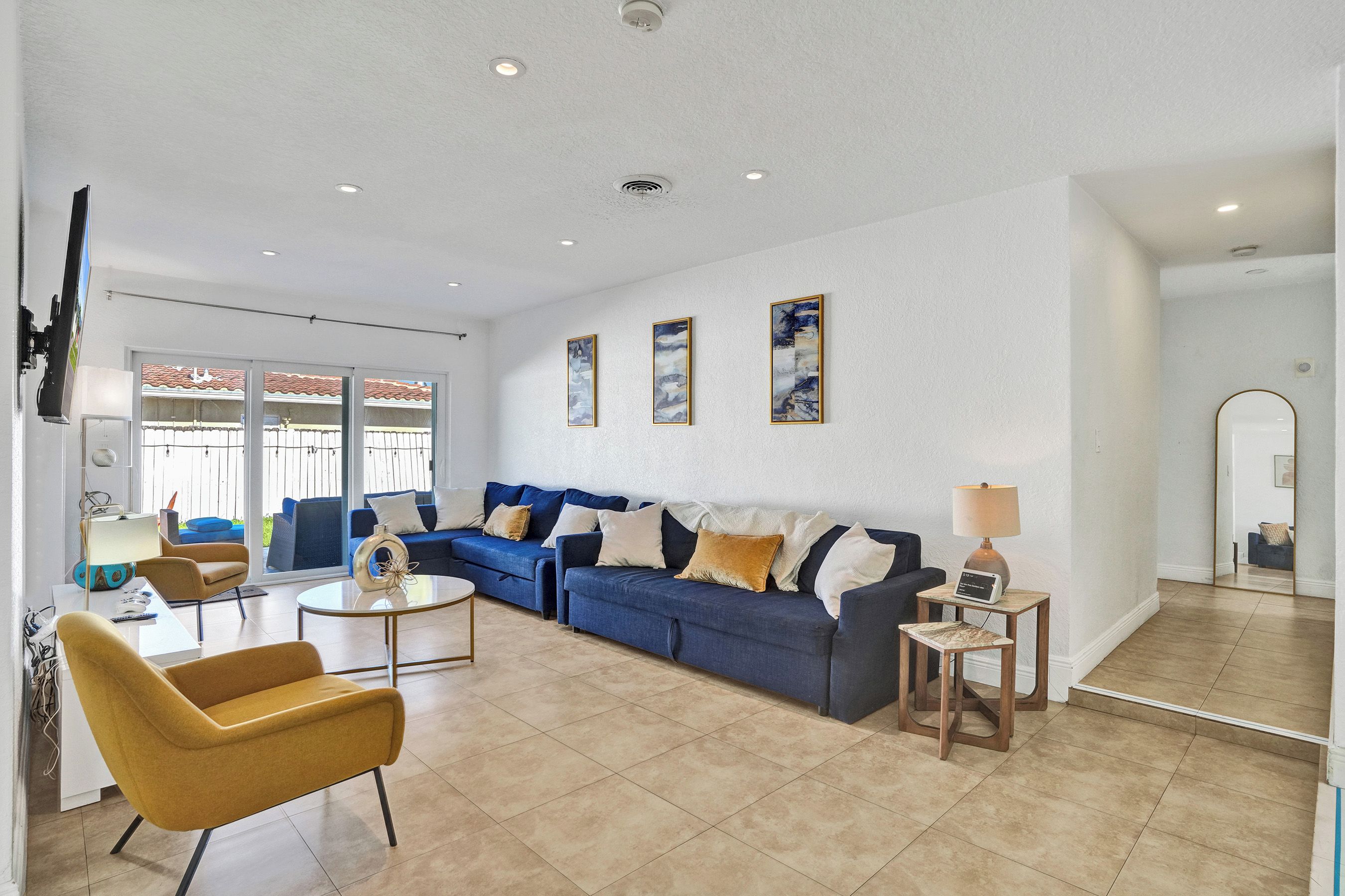 A spacious living room with a blue sofa, a yellow armchair, and a coffee table. There are three wall-mounted artworks and a lamp on a side table.