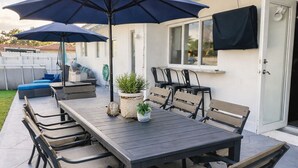 An outdoor patio with a rectangular table and multiple chairs, surrounded by potted plants and a large umbrella. - ★Miami Spacious Retreat 10 ninutes to Ocean • BBQ Pool Firepit• Sleeps 20+★ (North Miami Beach)