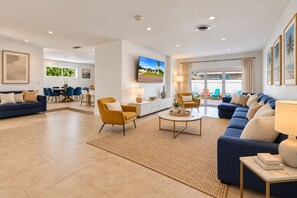 A spacious living room with a blue sofa, a yellow armchair, and a coffee table. There is a TV mounted on the wall, and a dining area with chairs and a table. - ★Miami Spacious 4BR Retreat •10 min to beaches• BBQ Firepit• Backyard★ (North Miami Beach)