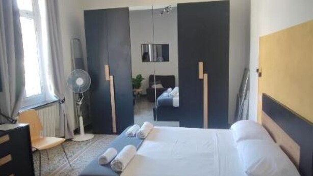 A hotel room with a bed, a desk, a chair, a standing fan, and a window with curtains. - Home Colors Apartment (Genova)