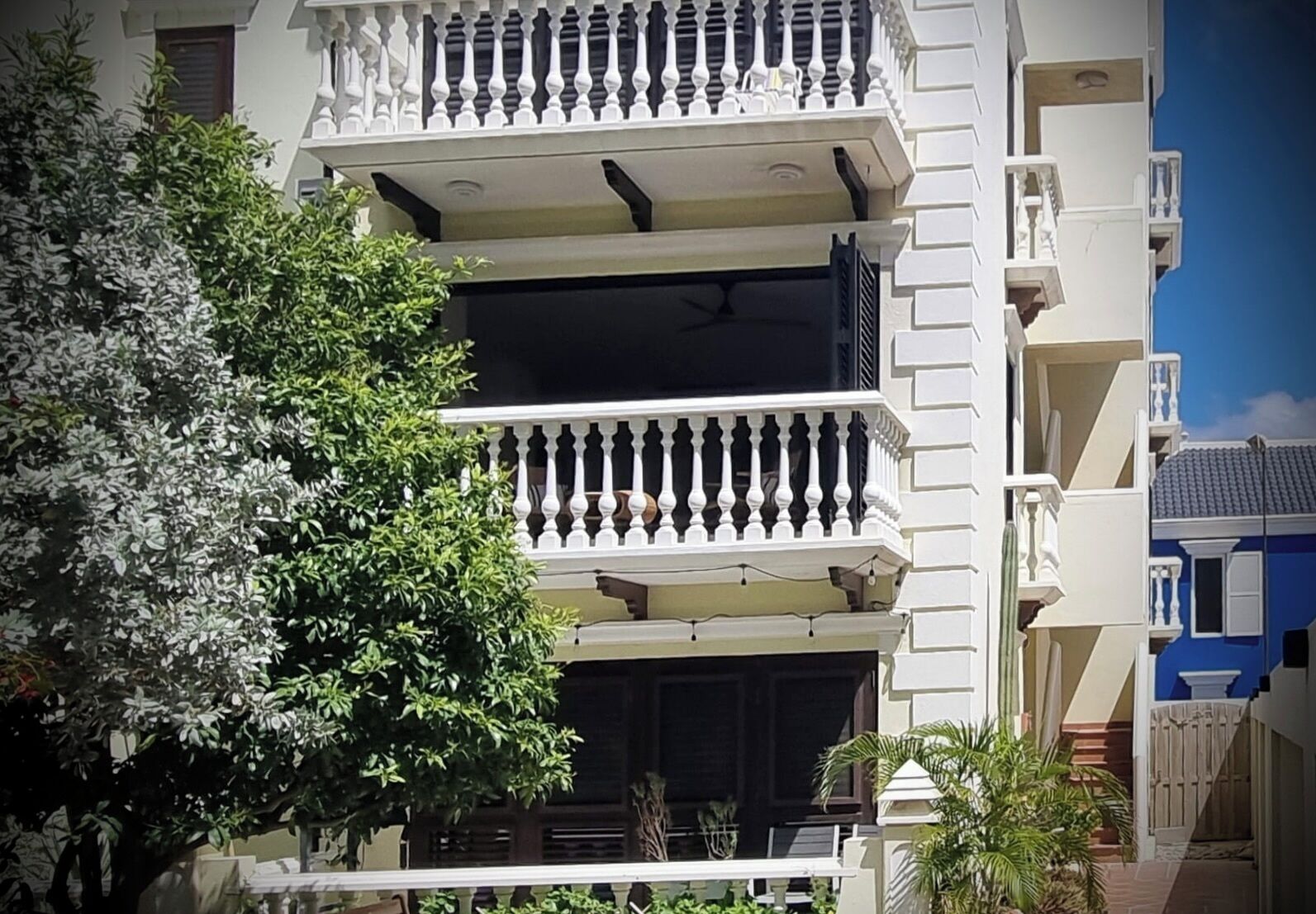 A multi-story building with balconies, a tree in the foreground, and a clear blue sky.