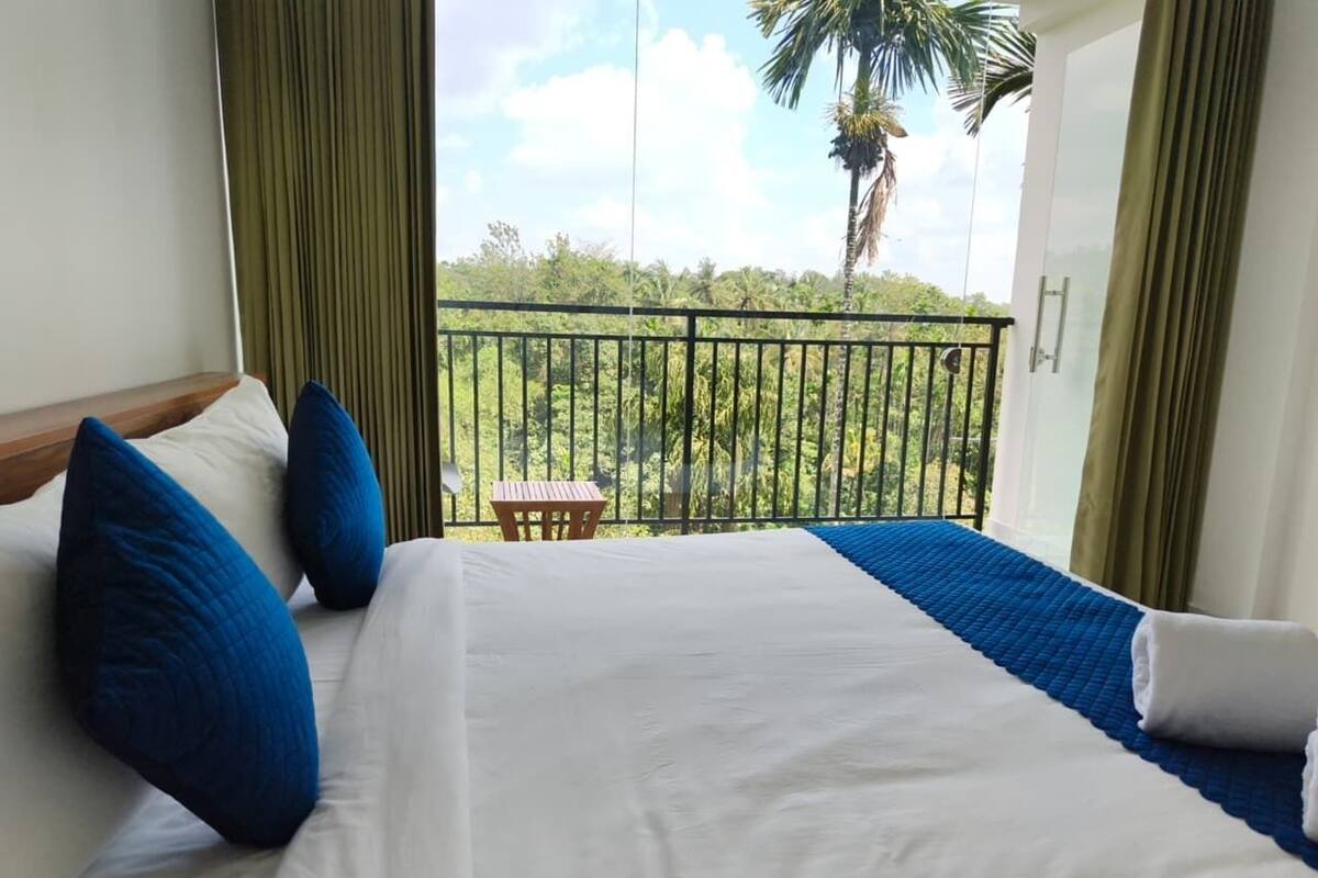 Una habitación de hotel con una cama grande, almohadas azules y vistas a la vegetación y una palmera afuera.