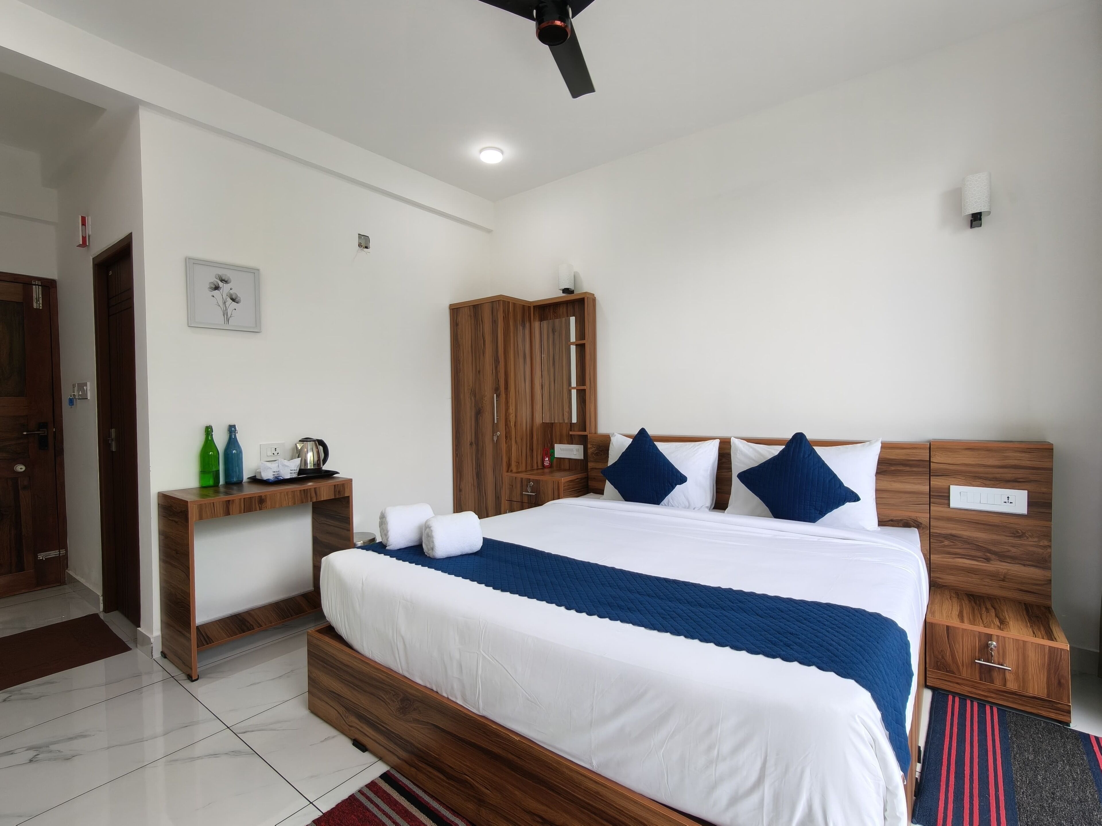 A bedroom with a wooden bed frame, a nightstand, a wooden wardrobe, and a desk with a kettle and bottles.