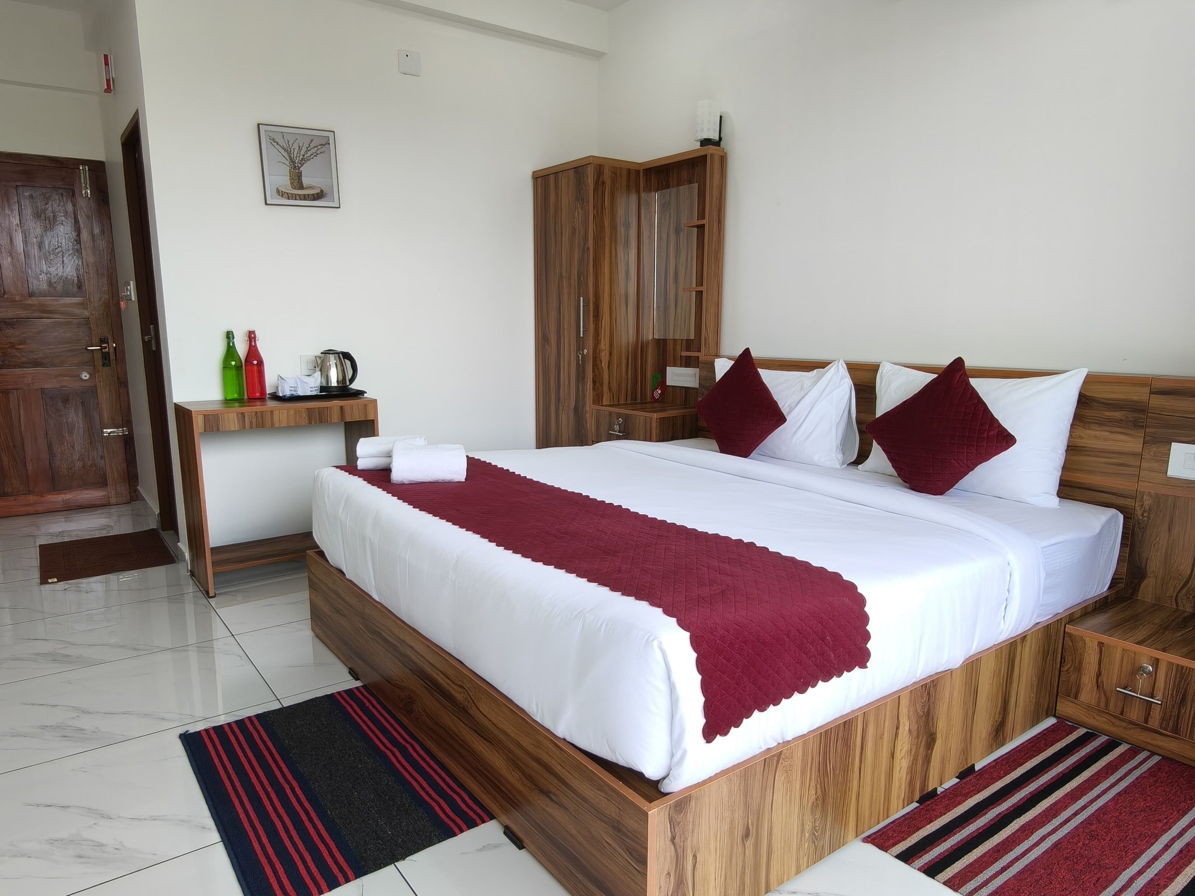 A hotel room with a large bed, wooden headboard, a bedside table with a kettle, and a nightstand.