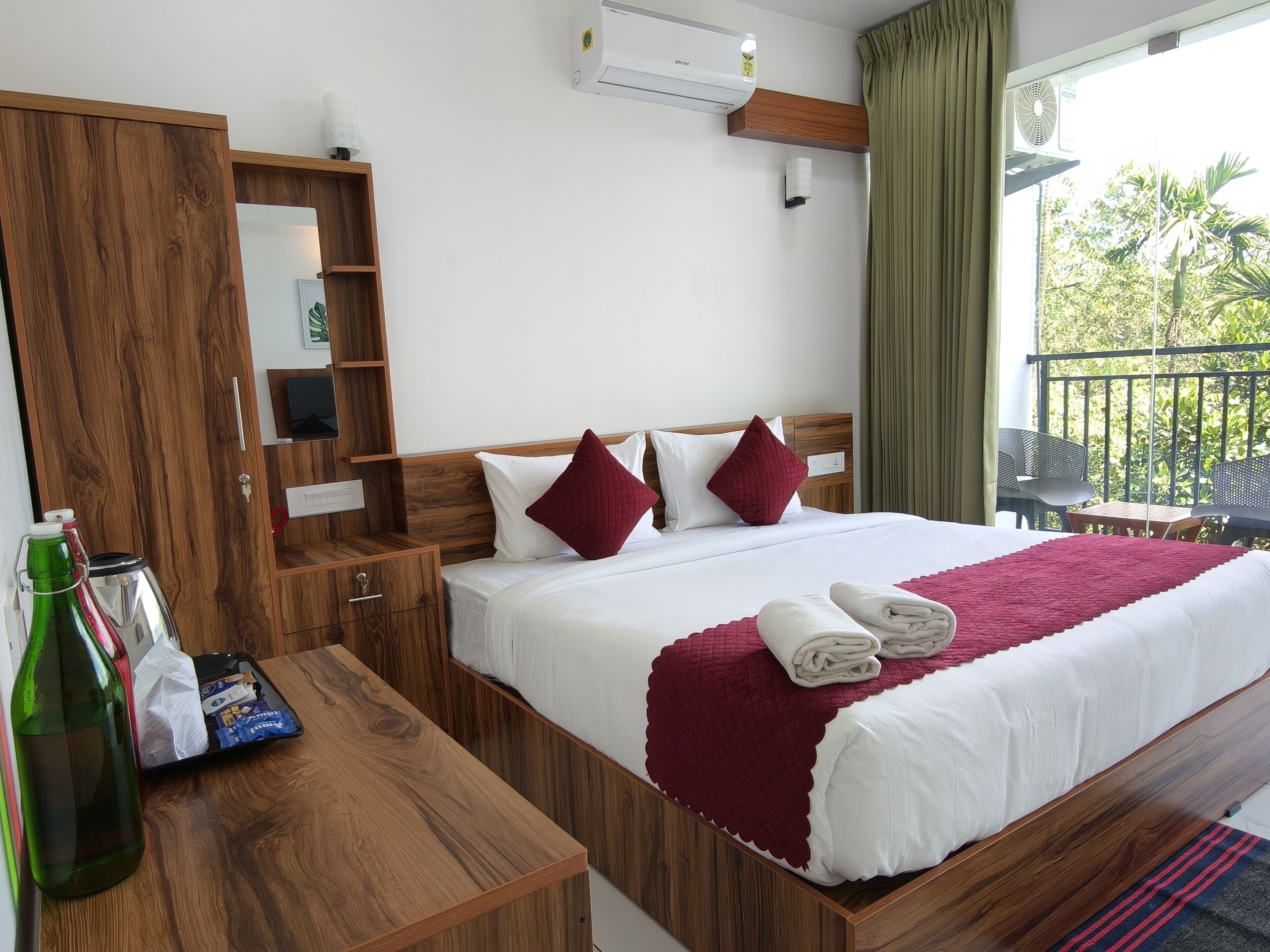 Habitación de hotel con una cama de madera, una mesita de noche, un espejo, un equipo de aire acondicionado y un balcón con mobiliario exterior.