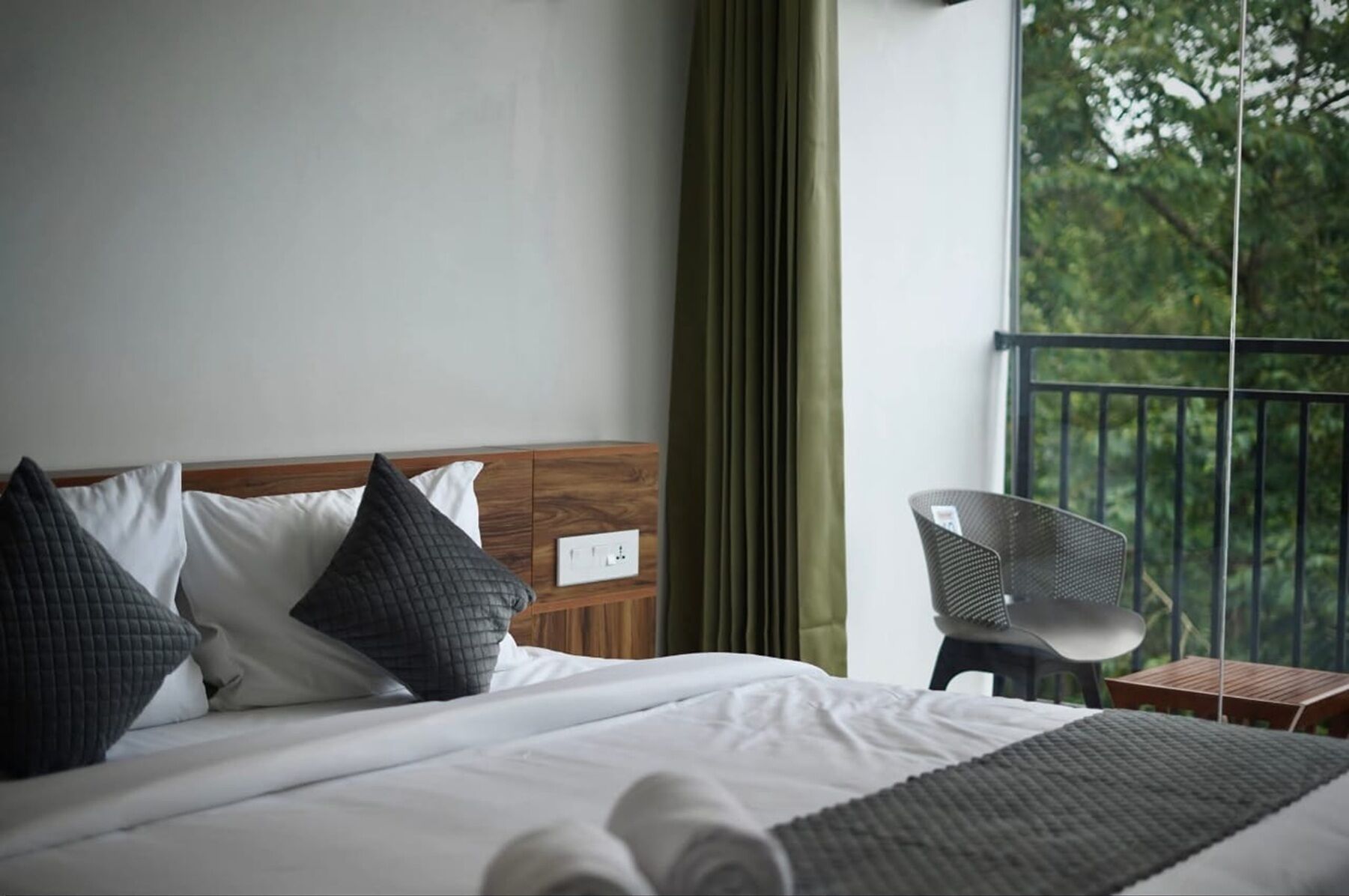 Habitación de hotel con cama, mesita de noche, una silla y vistas al exterior con vegetación.