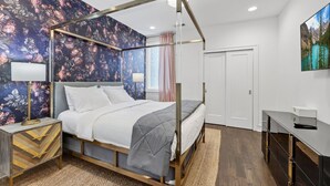 En seng med baldakin, blomstret tapet, et natbord med lampe, et skrivebord med lampe og en dør.