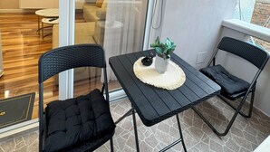 Parvekkeella on kaksi mustaa tuolia, pieni pöytä, jossa on kasvi, ja sieltä avautuu näkymä moderniin sisustukseen, jossa on myös televisio.