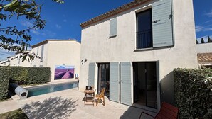 Une maison à deux étages avec une piscine, une terrasse dotée d’une table et de chaises, et une grande fenêtre avec des volets.