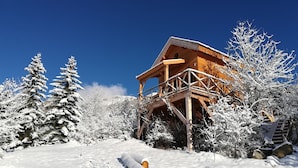 Un chalet en bois doté d’un balcon, entouré d’arbres couverts de neige et surplombant un ciel bleu clair.
