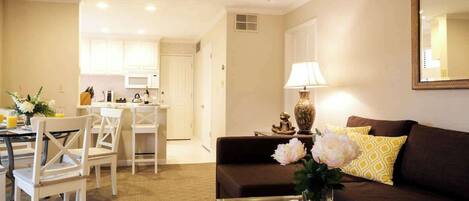 A living room with a brown sofa, a coffee table, and a dining area with white chairs and a kitchen in the background.