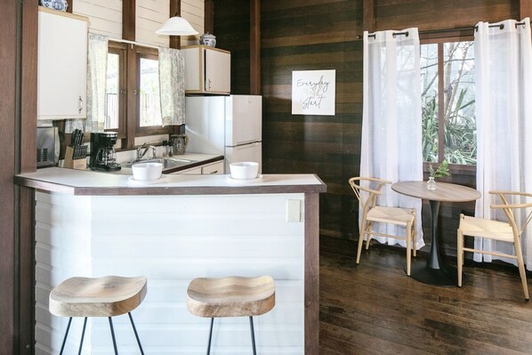 A kitchen with a central island, two bar stools, a dining table with chairs, and a window with curtains. - Oceanview Cottage Walk to Beach & Restaurants (Cruz Bay)