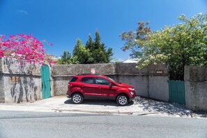 A red SUV parked in front of a concrete wall with a green gate, a clear blue sky, and trees in the background. - Oceanview Cottage Walk to Beach & Restaurants (Cruz Bay)