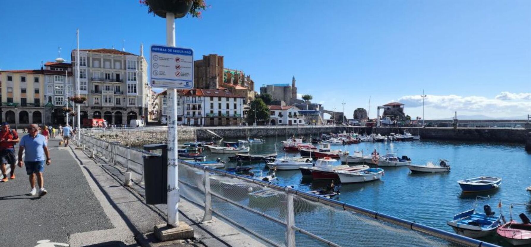 A waterfront promenade with boats docked, a pedestrian walkway, and historic buildings in the background.