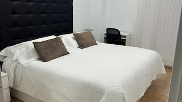 Ein ordentlich bezogenes Bett mit weißer Steudecke und zwei braunen Kissen, ein schwarzes, möbliertes Kopfteil, ein Nachttisch mit Lampe sowie ein Fenster mit vertikalen Rollläden.