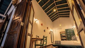 Una habitación con techo alto, vigas de madera, una cama, una silla, una mesa y luces fijadas a la pared.