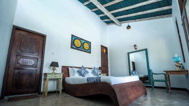 Una habitación con una cama de madera, una mesita con una lámpara, una silla, un espejo y una puerta.