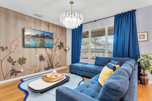 A modern living room with a blue sofa, a flat-screen TV mounted on the wall, a wooden floor, and a large window with blue curtains. - Castle Bunks & Viking Ship, Pool+Spa, 8 Min Disney (Kissimmee)