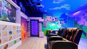 Ein Zimmer mit einer Wand im Minecraft-Design, einer Spielkonsole und einer Reihe von Gaming-Stühlen.
