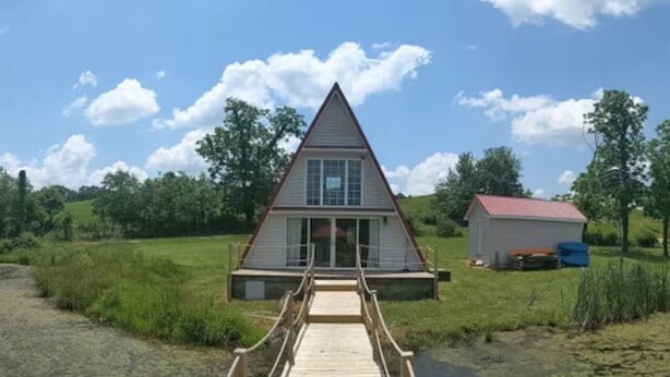 Un bâtiment triangulaire avec une terrasse en bois qui y mène, entouré de verdure et d’un petit abri de jardin.