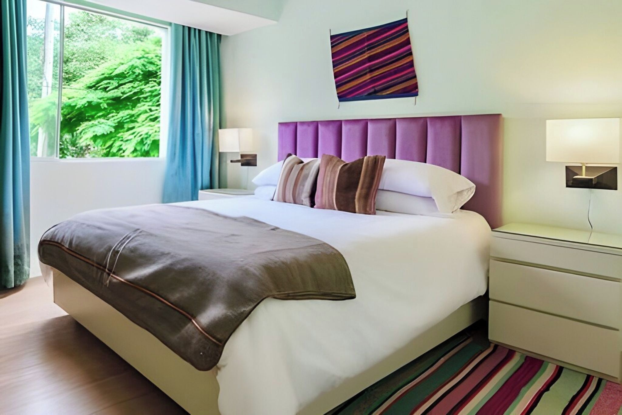 A modern hotel room with a large bed, a headboard, bedside tables, and a view of greenery outside.