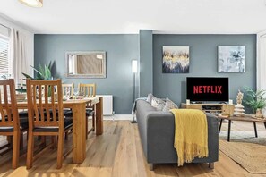 Olohuoneessa on harmaa sohva, puinen ruokapöytä ja tuolit, televisio, jossa näkyy Netflix, sekä taideteoksia seinillä.