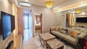 A modern living room with a large flat-screen TV, a chandelier, a sofa with cushions, a coffee table, and a TV stand. - Apartment Lincoln Podomoro Delipark (Medan)