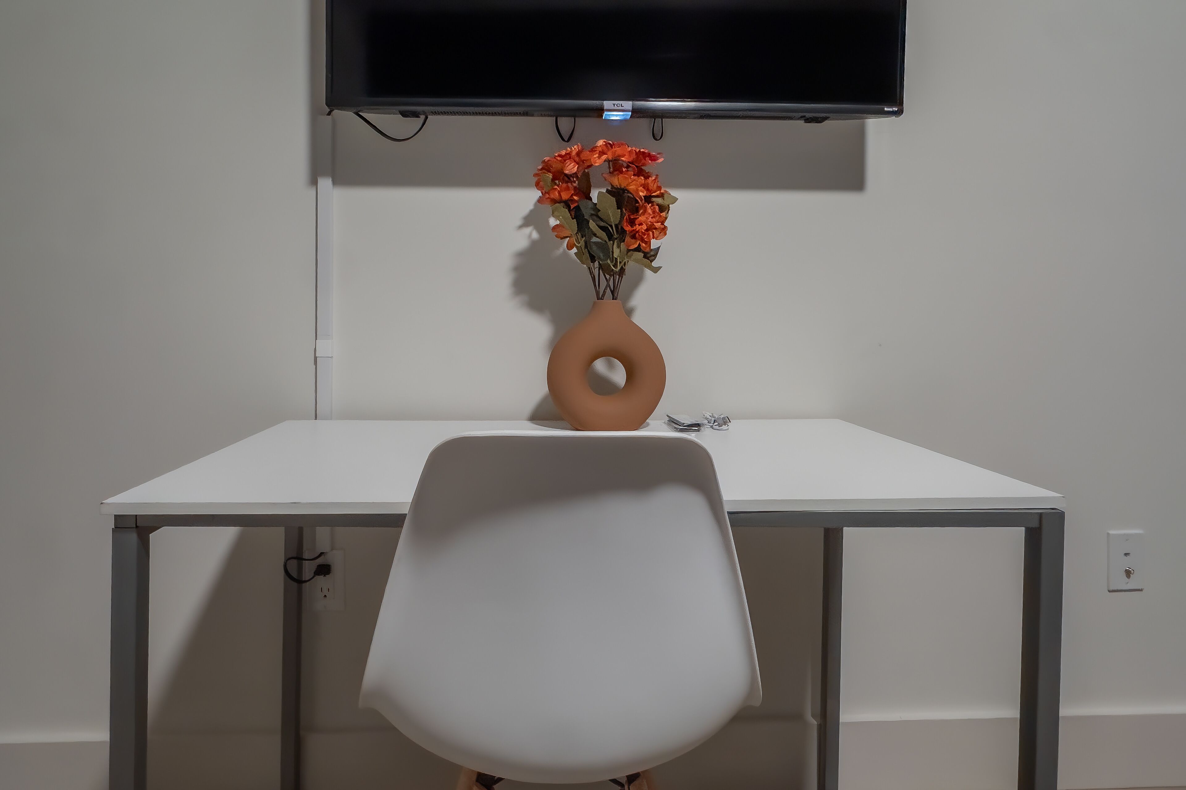 A white desk with a television mounted above it, a vase with orange flowers, and a white chair in front.